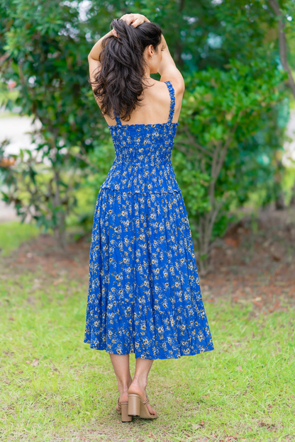 Woman in a blue floral dress standing outdoors with greenery in the background
