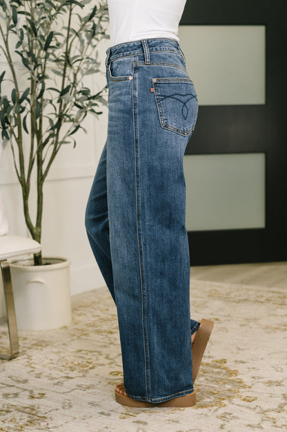Person wearing blue jeans standing indoors with a plant and chair in the background