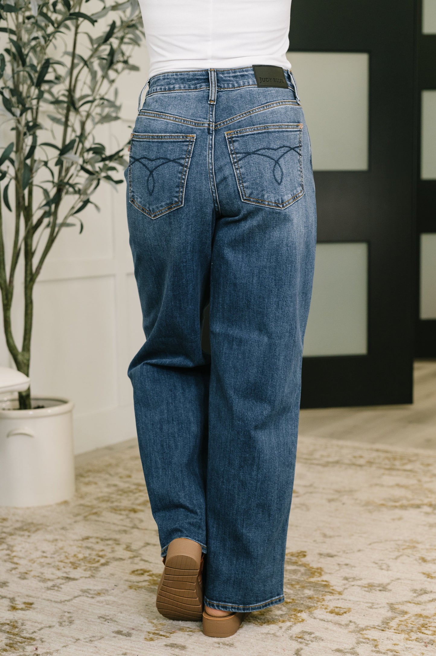 Person wearing blue jeans standing in a room with a plant and decorative wall.