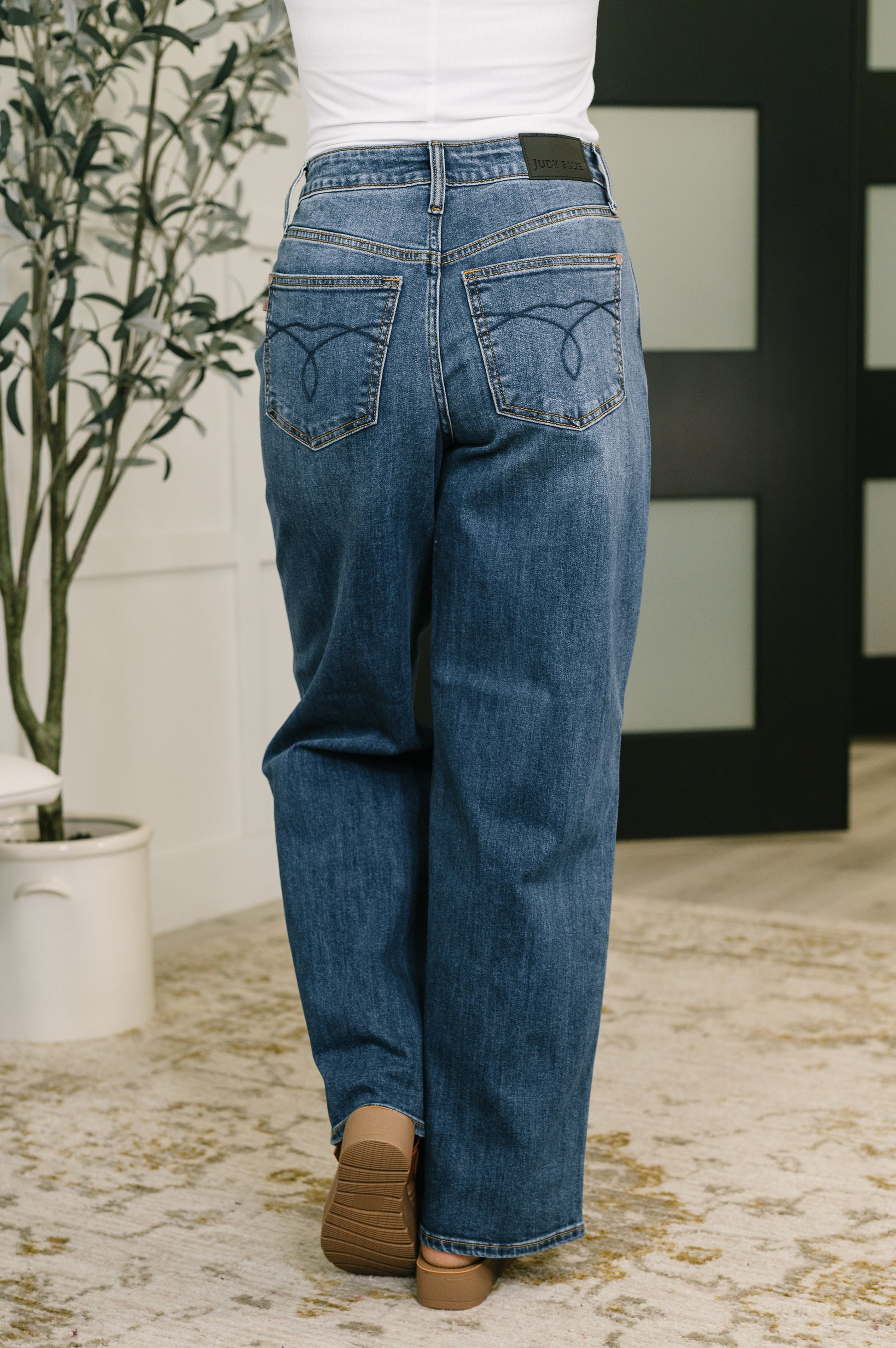 Person wearing blue jeans standing in a room with a plant and decorative wall.