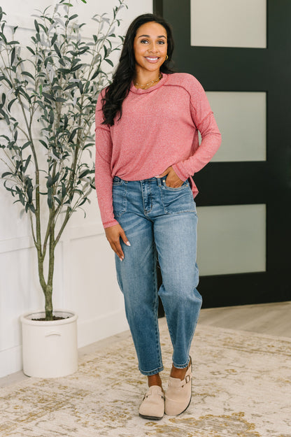 Woman wearing a pink sweater and blue jeans standing indoors with a plant and wall decor in the background.