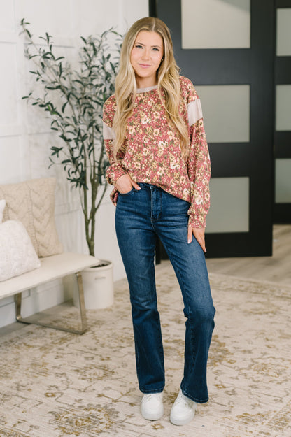 A woman stands in a bright indoor room wearing a floral long-sleeve top with sheer shoulder panels, dark blue jeans, and white sneakers.