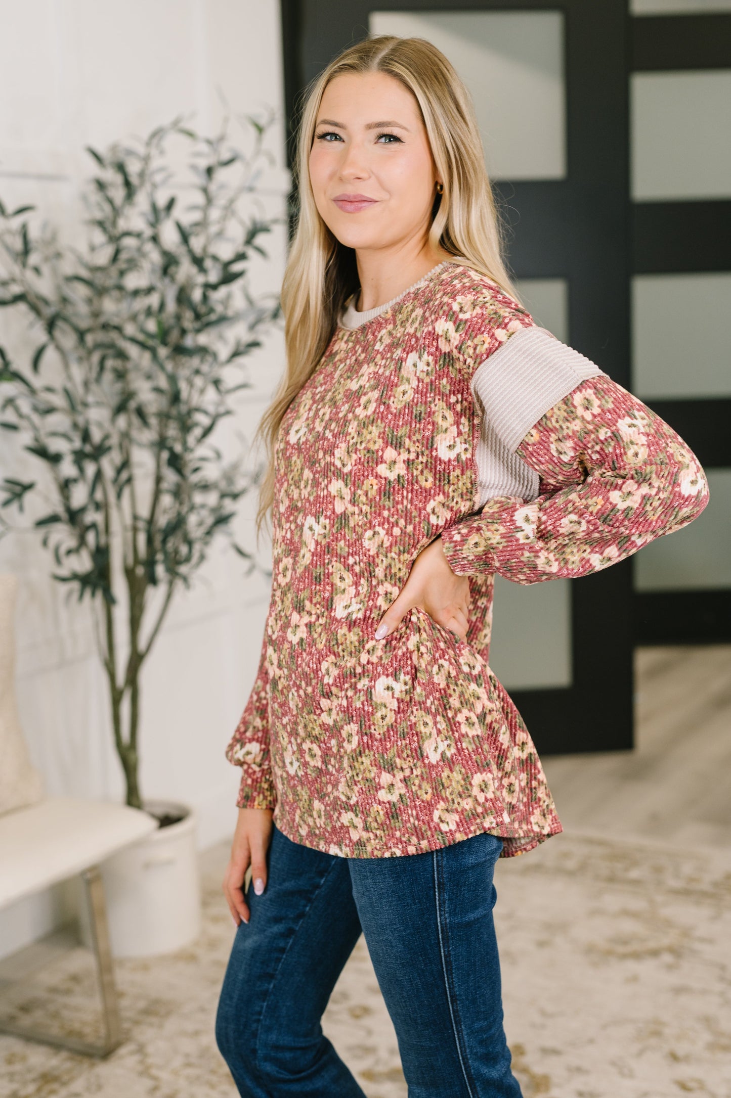 A woman modeling a long-sleeve floral top with a textured cream ribbed shoulder panel, paired with blue jeans in a modern indoor setting.