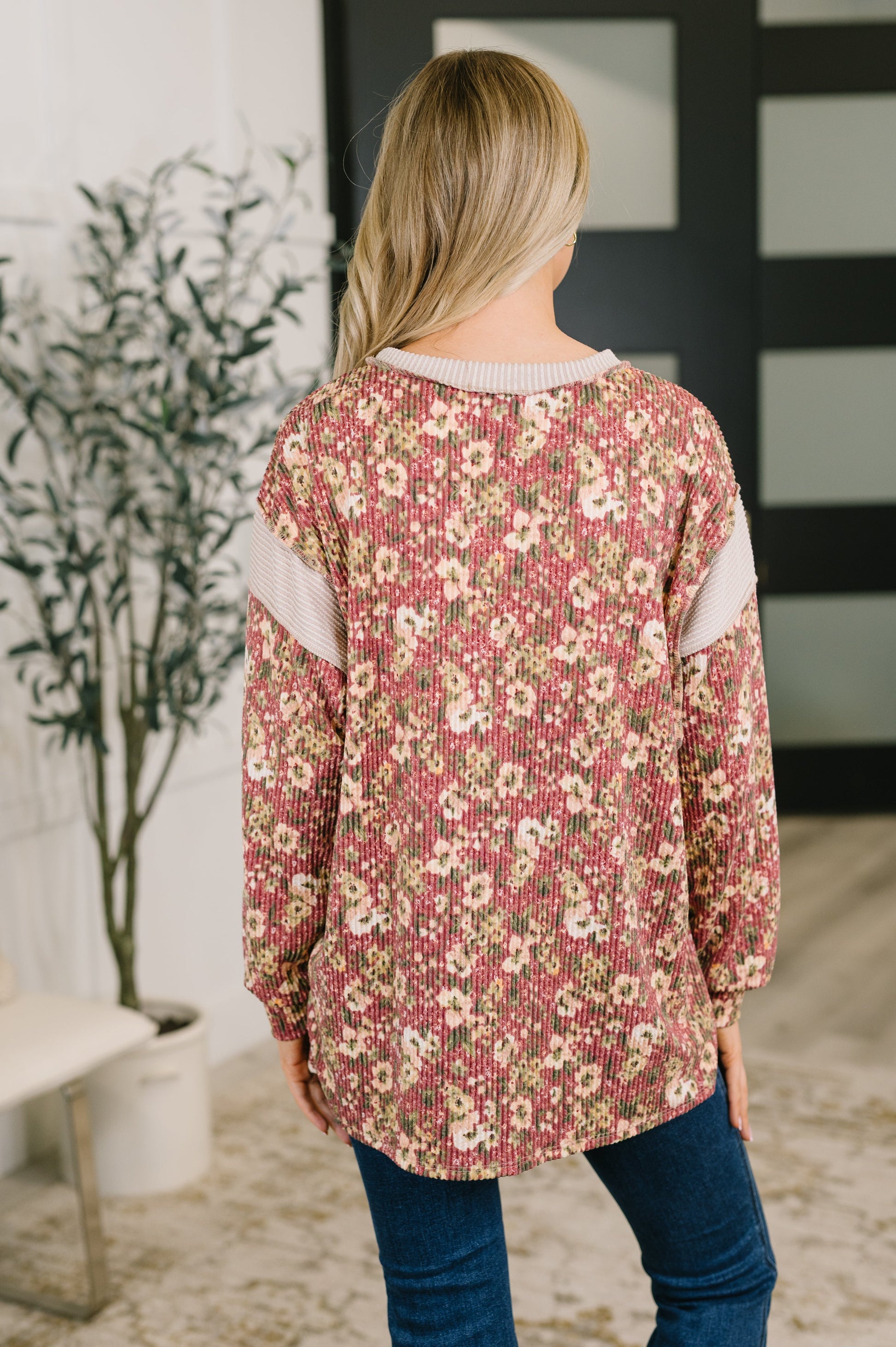 A woman viewed from behind wearing a long-sleeve floral knit top with pink, beige, and white tones, paired with blue jeans in an indoor setting.