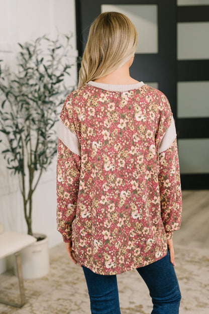 A woman viewed from behind wearing a long-sleeve floral knit top with pink, beige, and white tones, paired with blue jeans in an indoor setting.