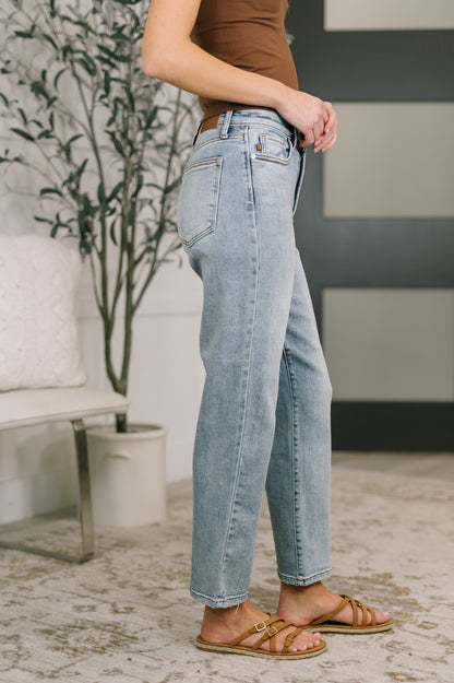 Person wearing light blue jeans and brown sandals in a room with a plant and furniture.