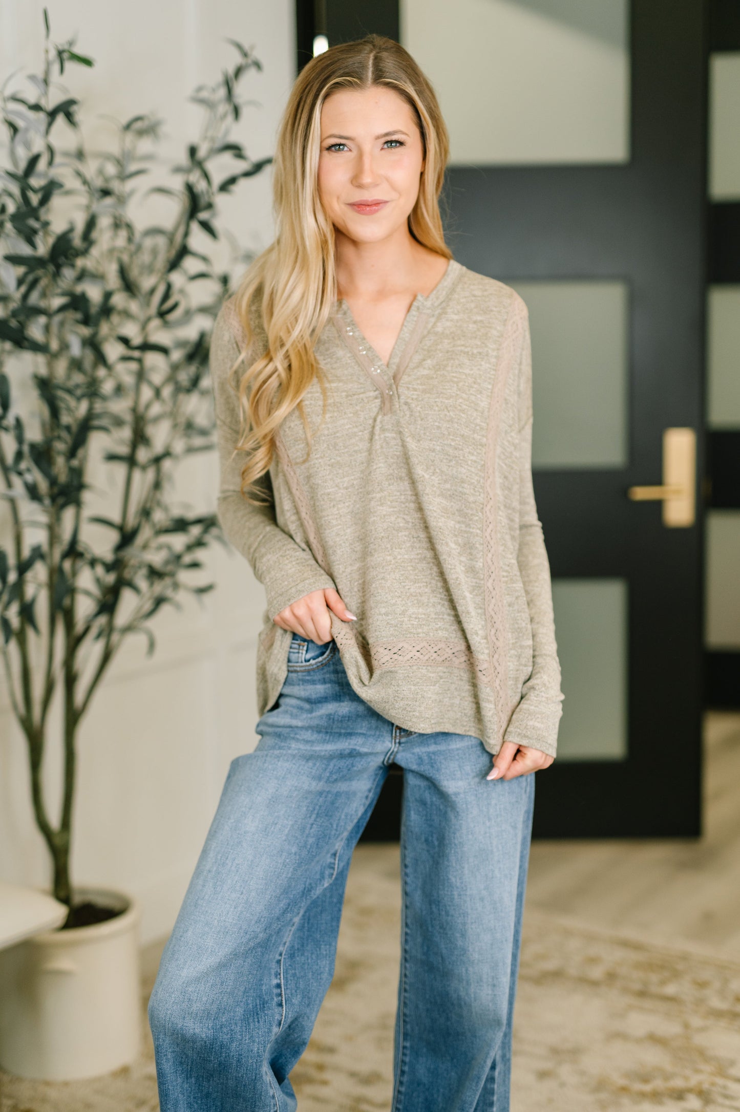 A woman modeling a light beige knit sweater with a split neckline and lace-up detail, paired with wide-leg blue jeans in a modern indoor setting with a potted plant and a black door in the background.