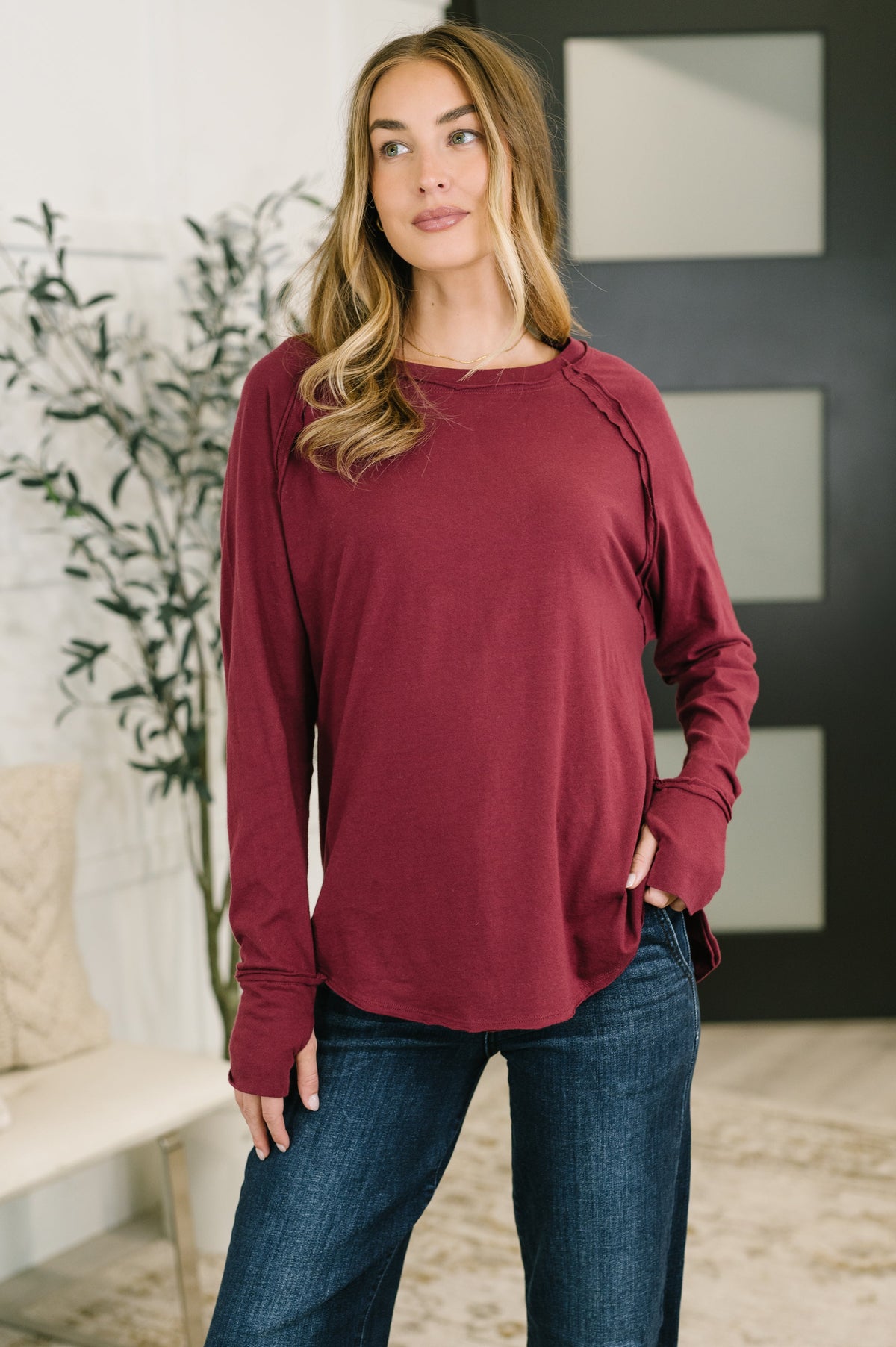A woman wearing a long-sleeve maroon top with ruched shoulder details and side slits, paired with dark blue jeans, standing in a modern indoor setting.
