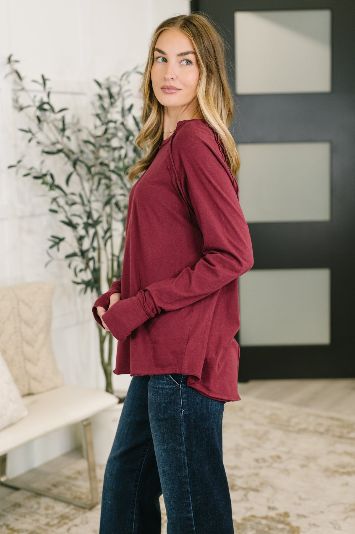 A woman modeling a long-sleeve maroon top with a relaxed, slightly oversized fit, paired with dark blue jeans in a modern indoor setting.