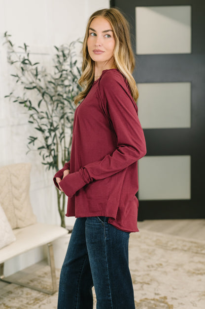 A woman modeling a long-sleeve maroon top with a relaxed, slightly oversized fit, paired with dark blue jeans in a modern indoor setting.