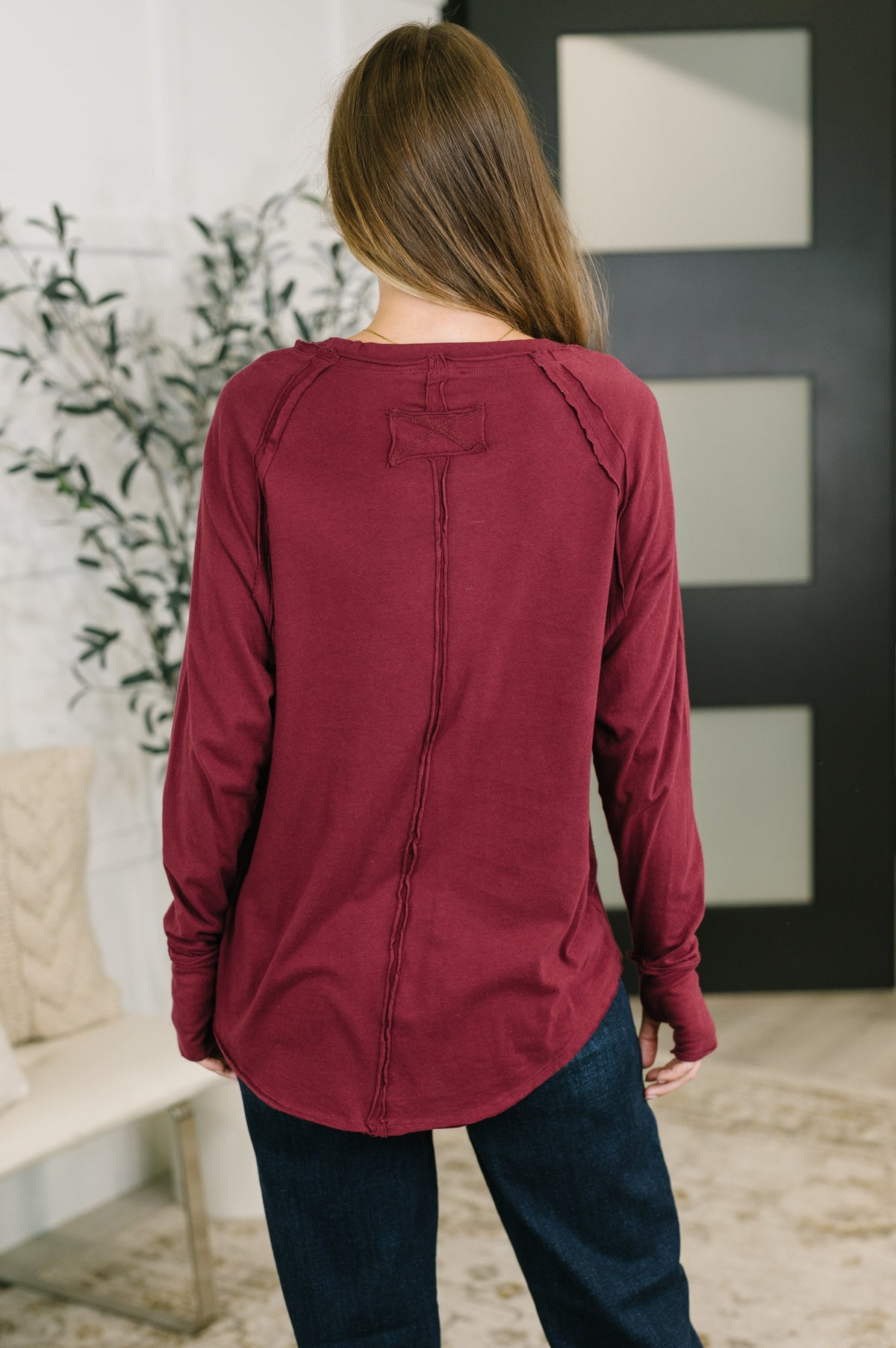 A woman viewed from behind wearing a long-sleeve maroon top with a decorative bow detail at the back neckline, paired with dark blue jeans in an indoor setting.