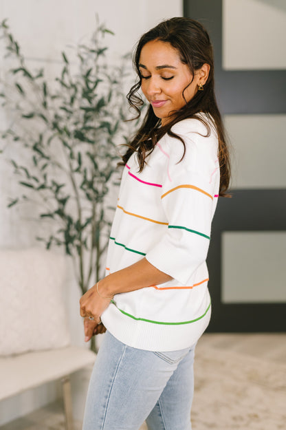 Woman wearing a white sweater with colorful stripes in a living room setting.