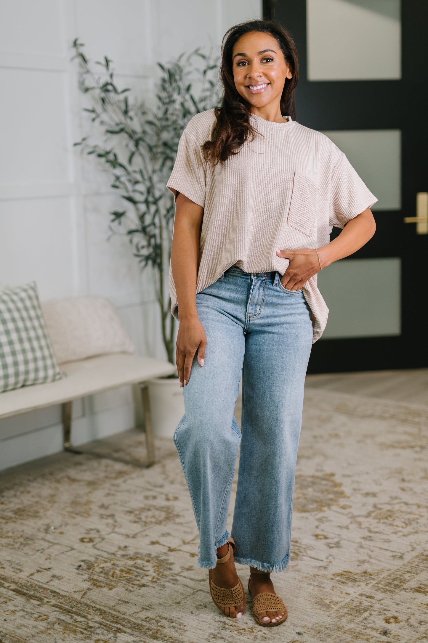 Woman wearing a beige top and light blue jeans standing in a room with a plant and bench.