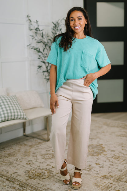 Woman wearing a turquoise top and beige pants standing in a room with a couch and plant.