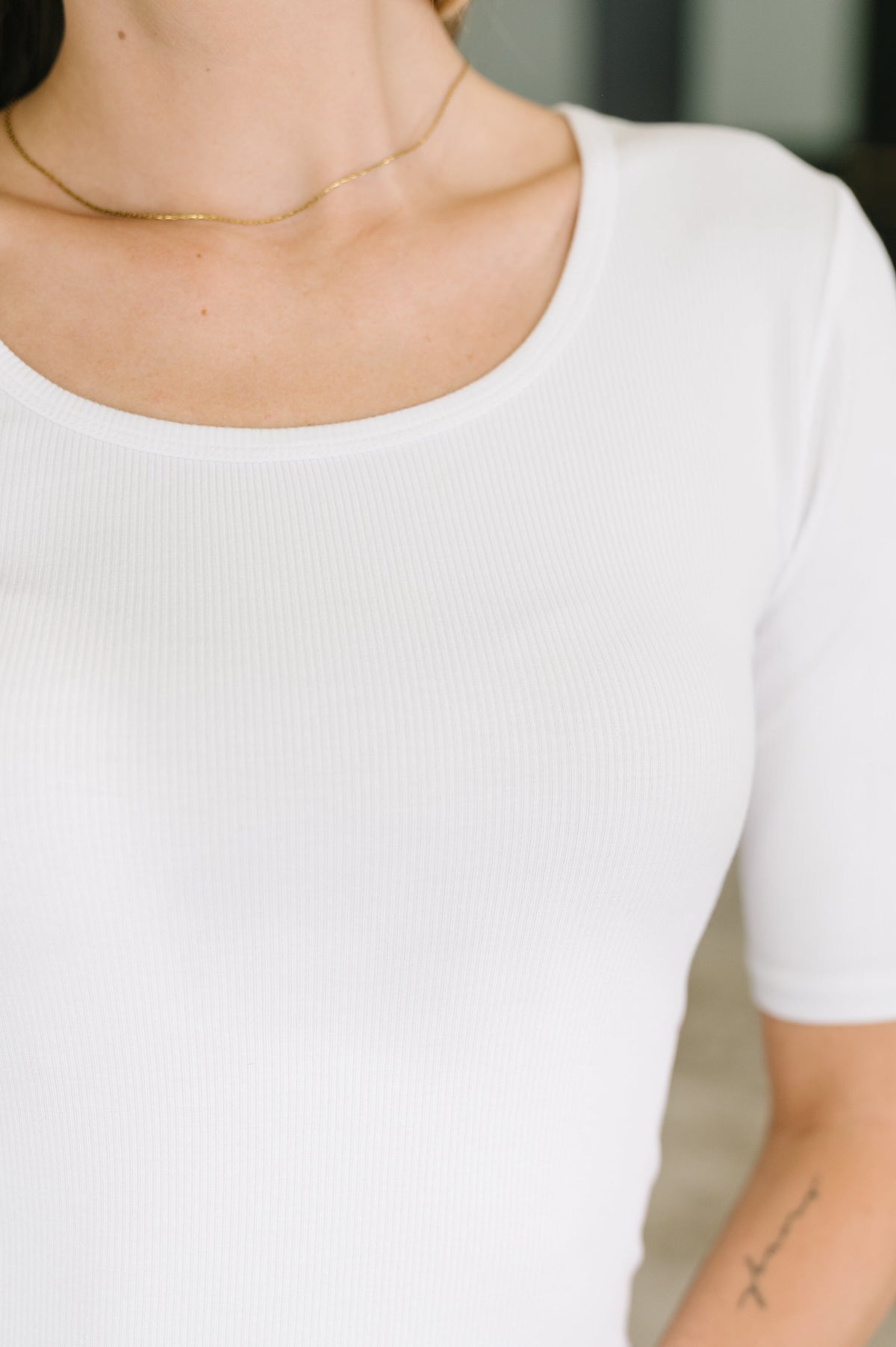 Close-up of a person wearing a white short-sleeve ribbed scoop-neck top, with a delicate gold chain necklace visible.