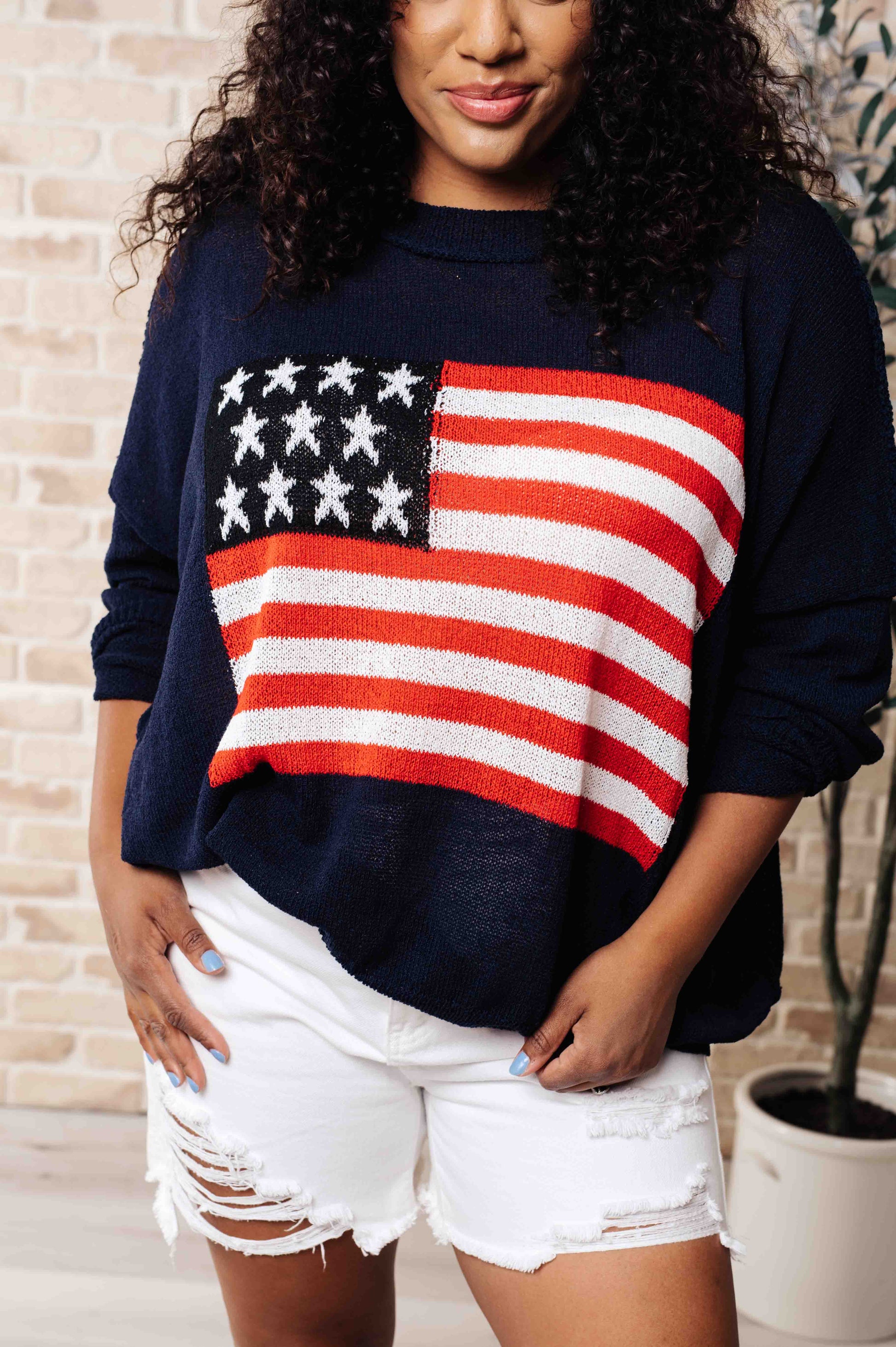 A woman wearing a navy blue lightweight pullover with a round neckline and a flag pattern on the front, paired with white high-waisted shorts.