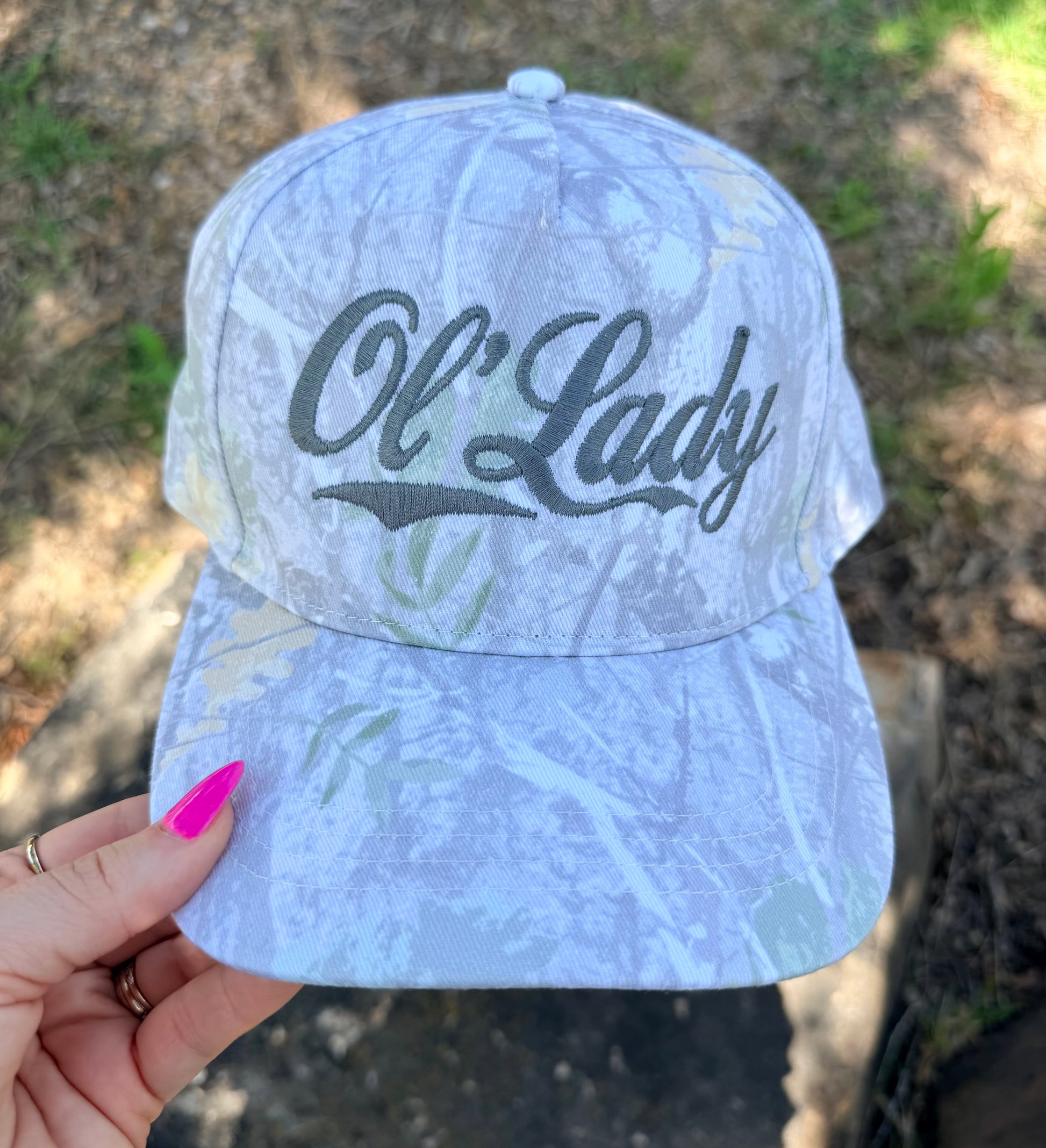 A light blue and white camouflage baseball cap with 'Ol' Lady' embroidered in black script on the front, held by a hand with bright pink fingernails against an outdoor background.