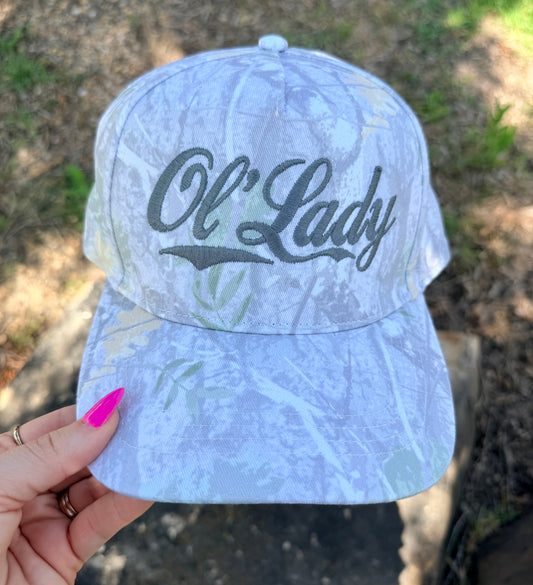 A light blue and white camouflage baseball cap with 'Ol' Lady' embroidered in black script on the front, held by a hand with bright pink fingernails against an outdoor background.