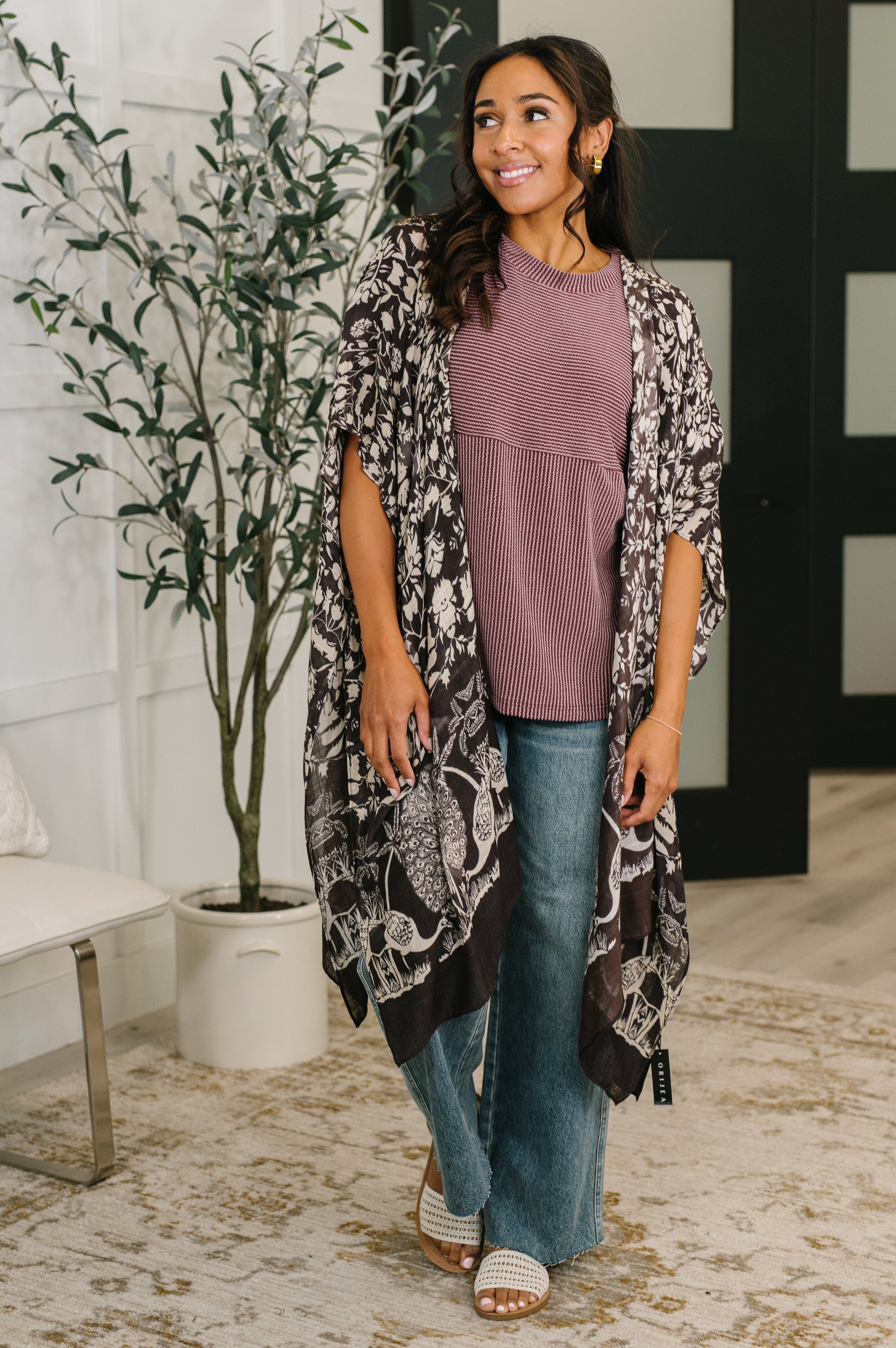 Woman wearing a floral kimono over a purple top and blue jeans, standing indoors with a plant and bench in the background.
