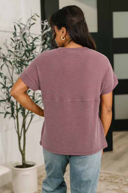 Person wearing a maroon ribbed top and light blue jeans standing indoors with a plant in the background.