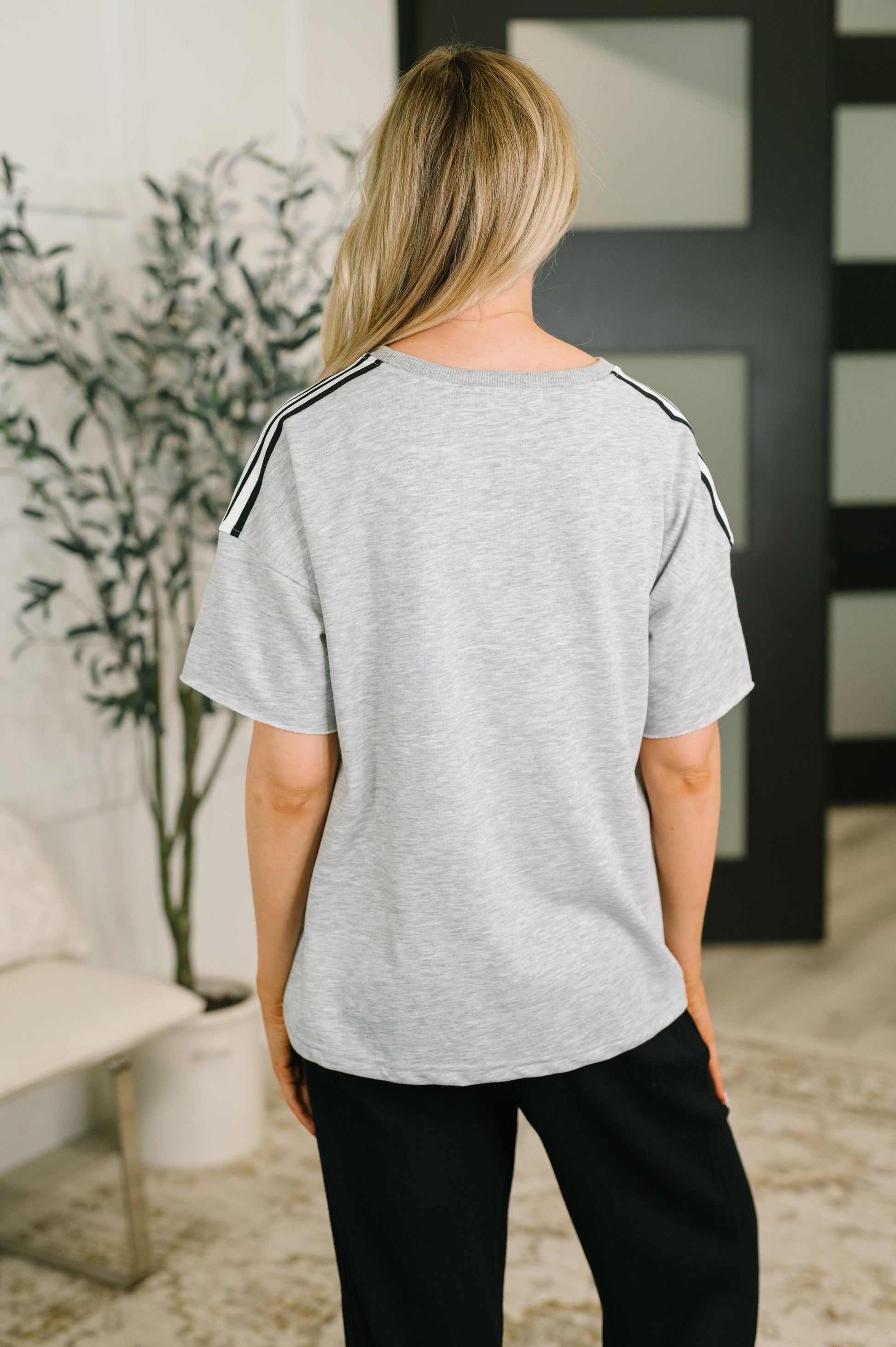 A woman shown from the back wearing a light heather gray short-sleeve top with black shoulder stripes, paired with black pants, in an indoor setting.