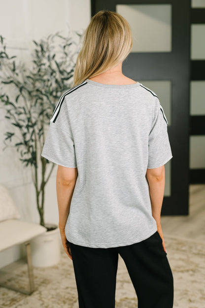 A woman shown from the back wearing a light heather gray short-sleeve top with black shoulder stripes, paired with black pants, in an indoor setting.