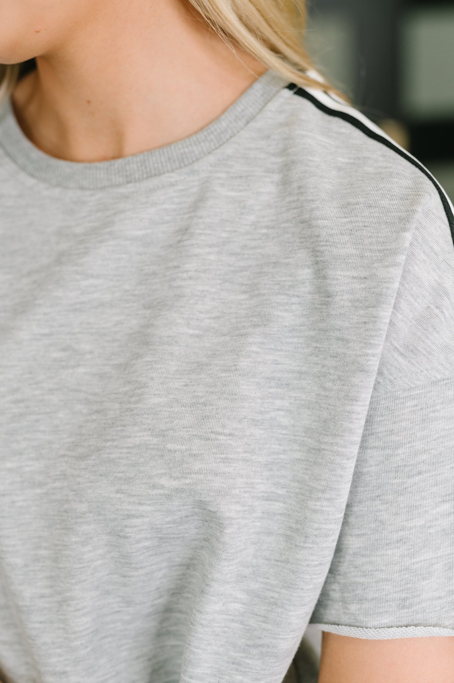 Close-up of a person wearing a light heather gray short-sleeve crewneck shirt with a thin black stripe detail along the shoulder.