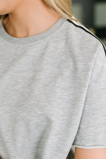 Close-up of a person wearing a light heather gray short-sleeve crewneck shirt with a thin black stripe detail along the shoulder.
