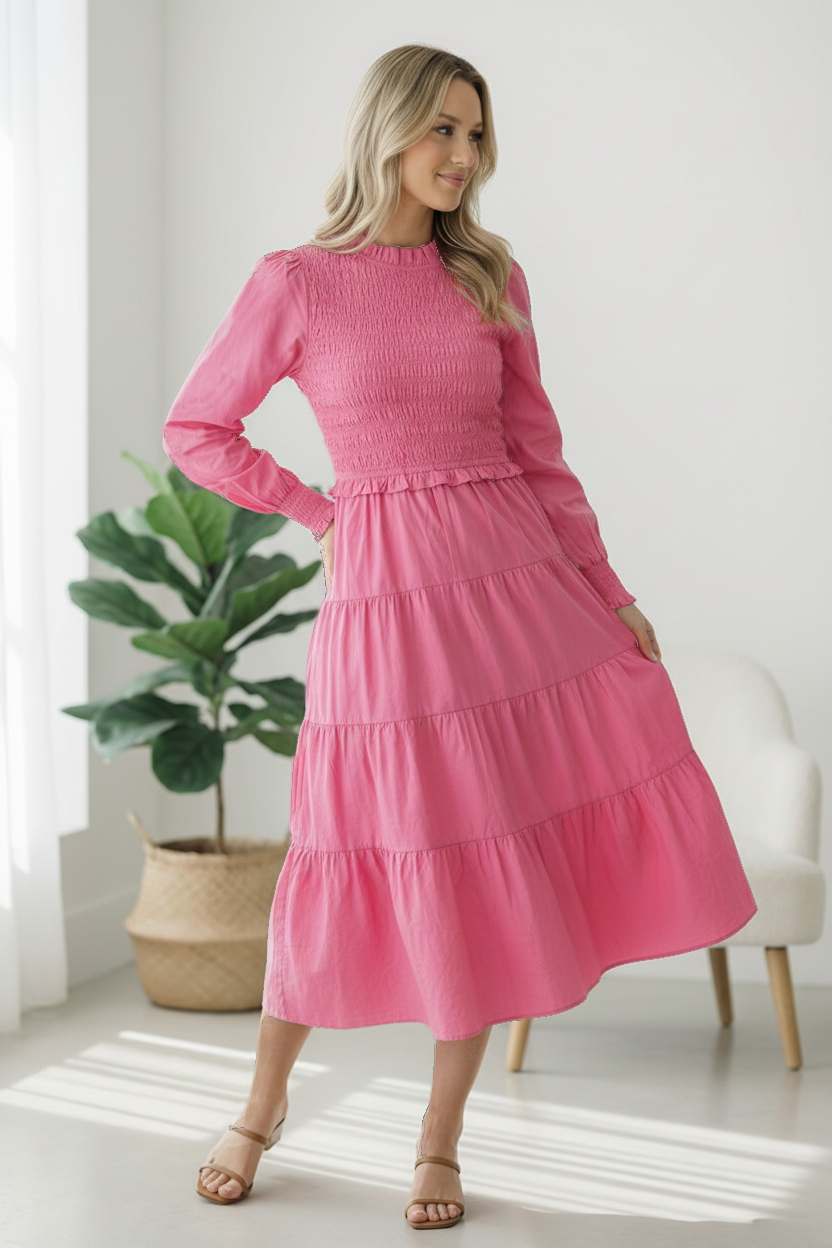 Woman wearing a pink dress standing in a bright room with a plant and chair.