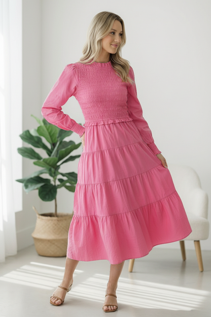 Woman wearing a pink dress standing in a bright room with a plant and chair.