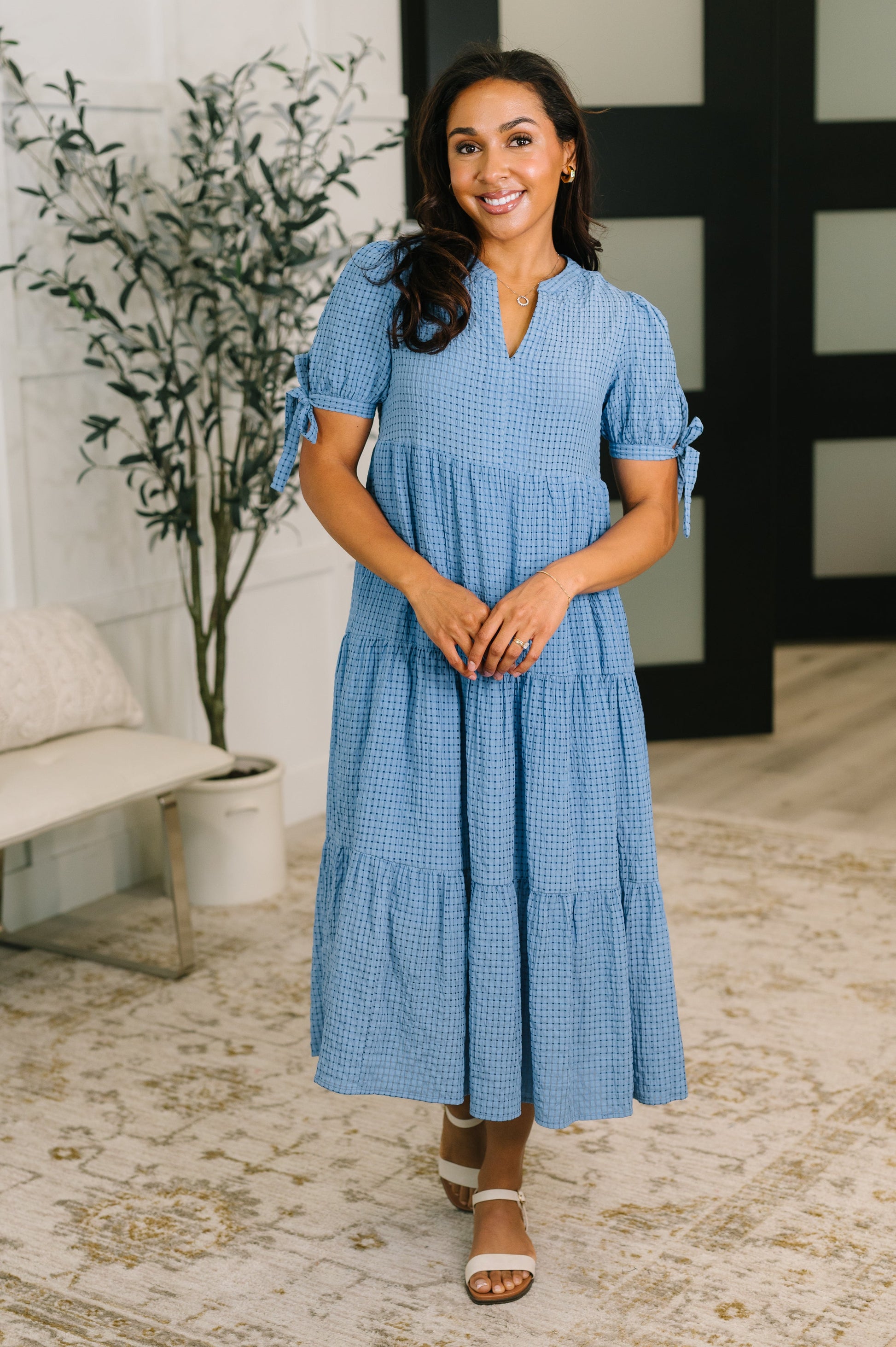 Woman wearing a blue checkered dress standing in a room with a plant and sofa.