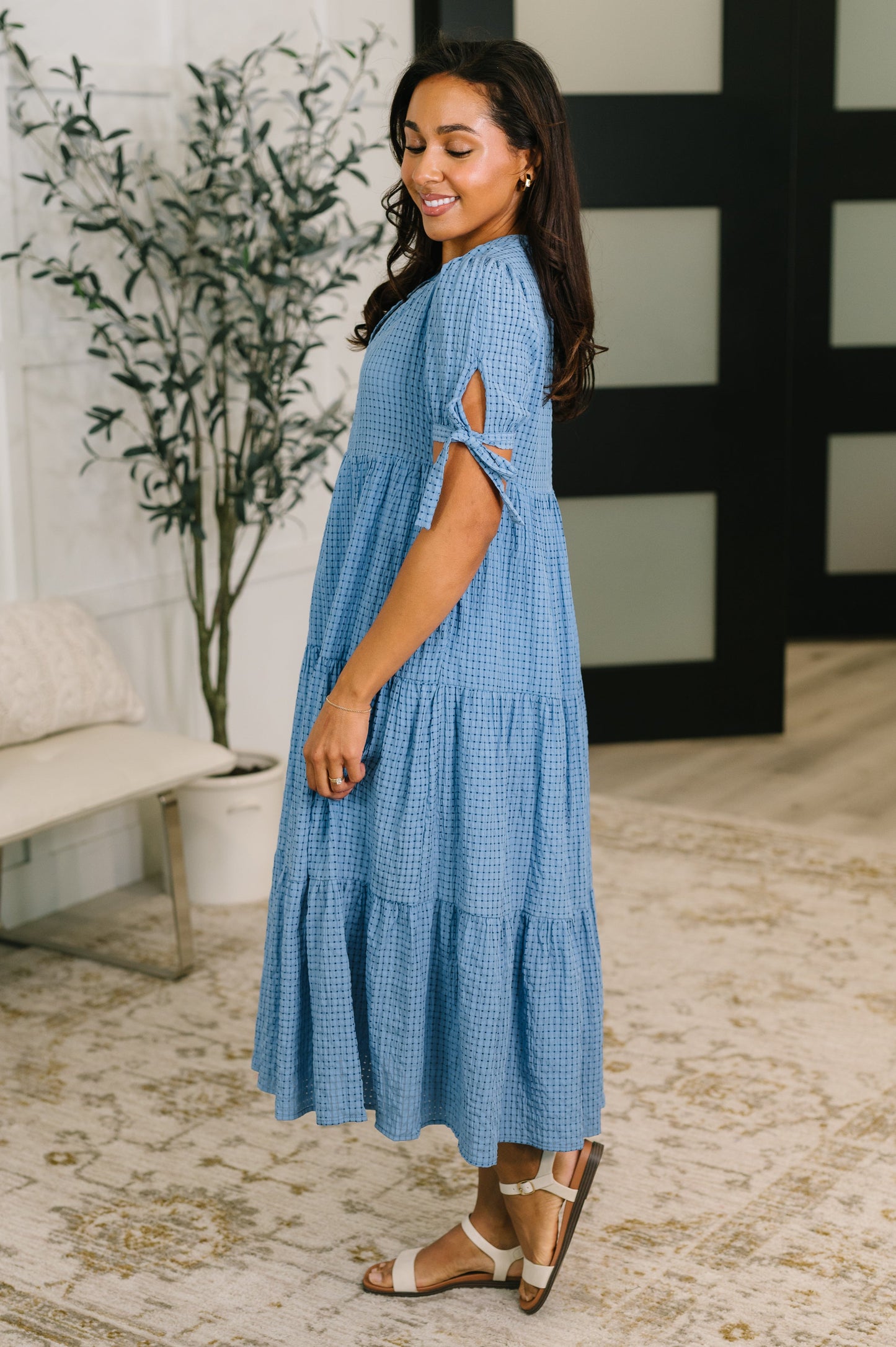 Woman wearing a blue dress standing in a room with a plant and decorative wall.