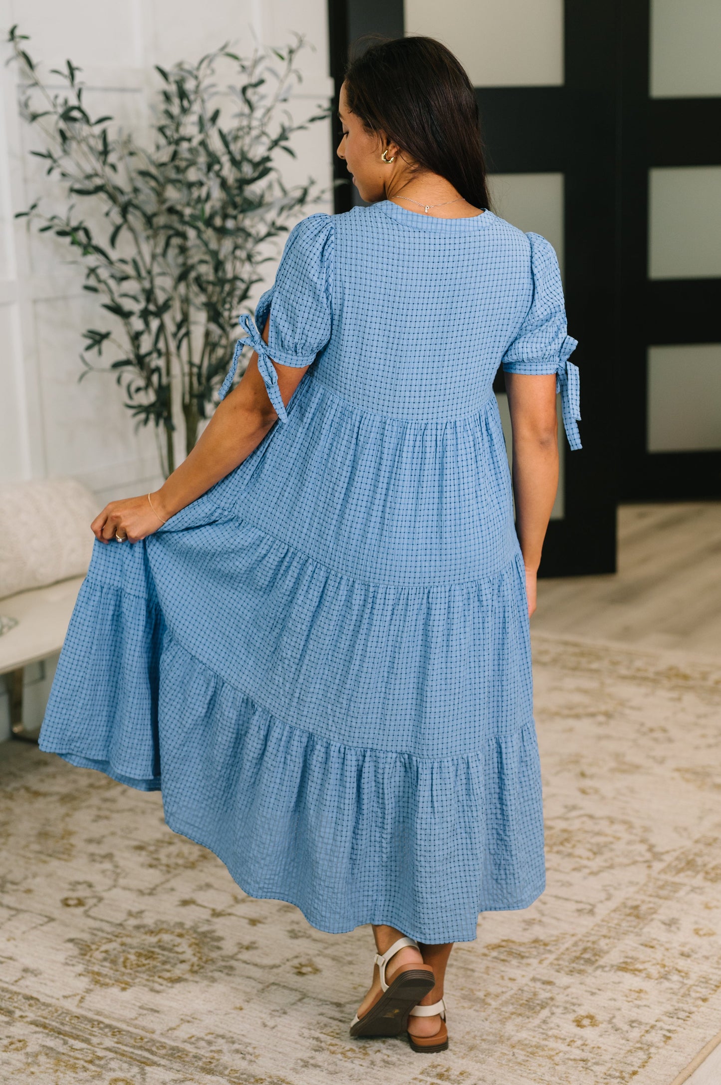 Woman wearing a blue checkered dress in an indoor setting with a plant and decorative wall.