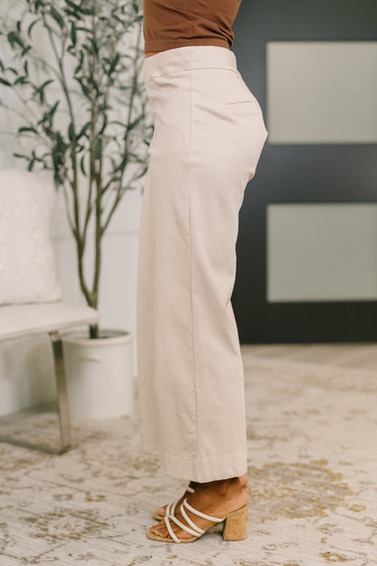 Person wearing beige pants and a brown top in an indoor setting with a plant and sofa.