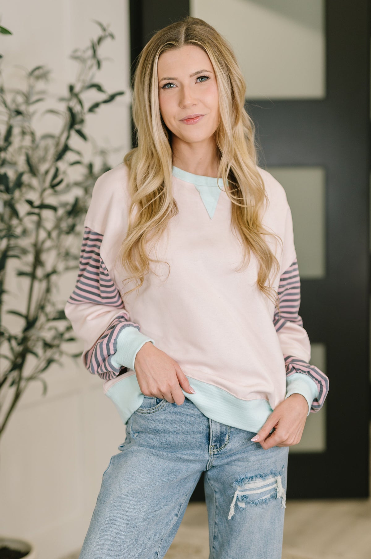A woman modeling a pastel crewneck sweatshirt with light pink, mint green, and striped sleeves, paired with light-wash distressed jeans.