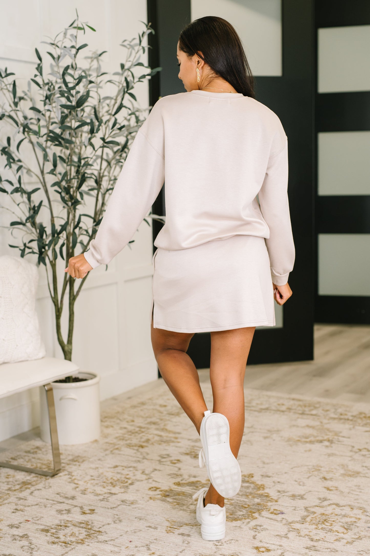 Woman in a white outfit walking in a modern indoor setting with a plant and white furniture.