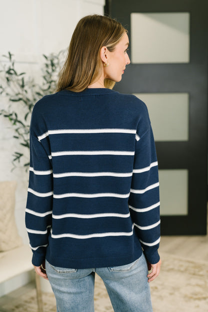 A woman viewed from the back wearing a navy blue knit sweater with horizontal white stripes and light blue jeans, standing indoors near a modern dark door and light-colored wall.