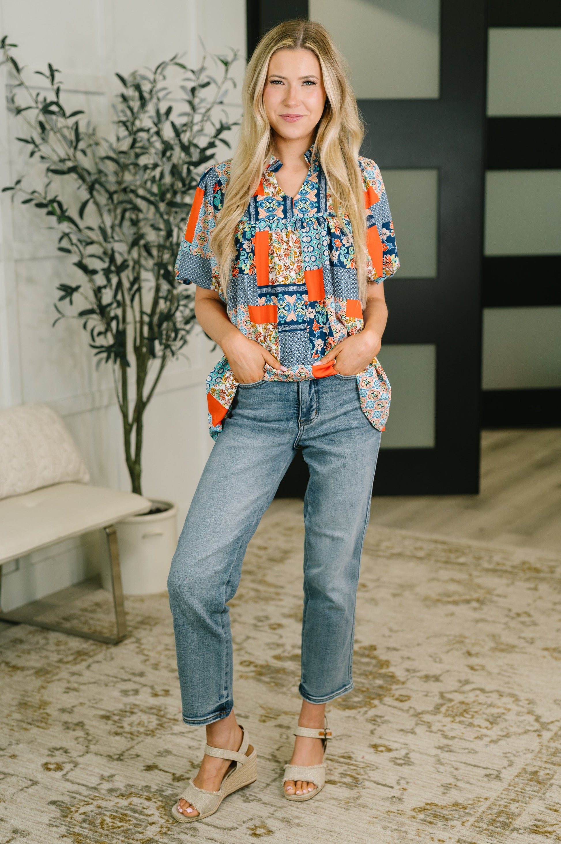 A woman stands indoors wearing a short-sleeve button-up shirt with a colorful patchwork-style print in blue, orange, and white, paired with light blue jeans and beige wedge sandals.