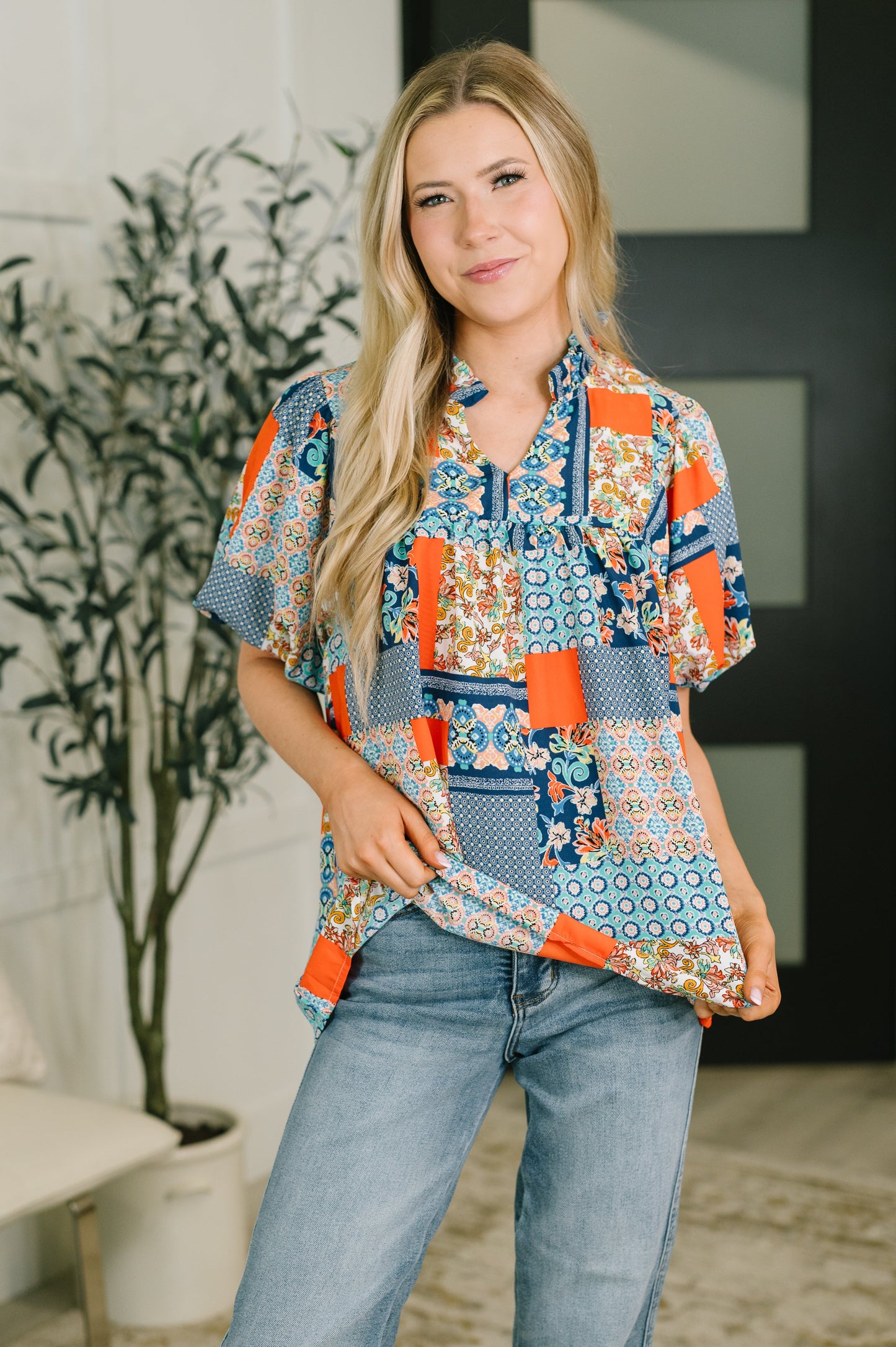 A woman modeling a short-sleeve button-up shirt with a colorful patchwork print in blue, orange, and white, paired with light-wash blue jeans in an indoor setting.
