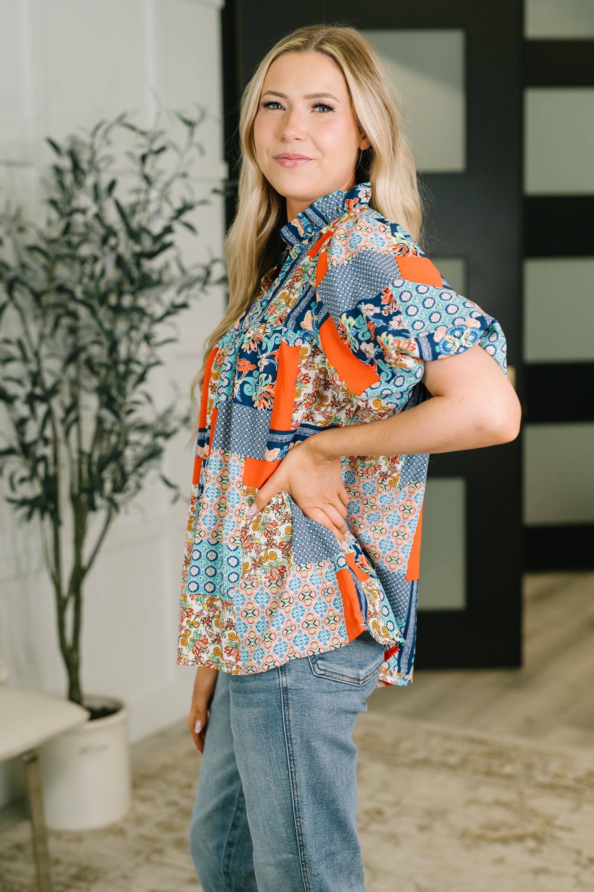 A woman modeling a short-sleeve button-up shirt with a colorful patchwork print of blue, orange, and beige patterns, paired with blue jeans in an indoor setting.