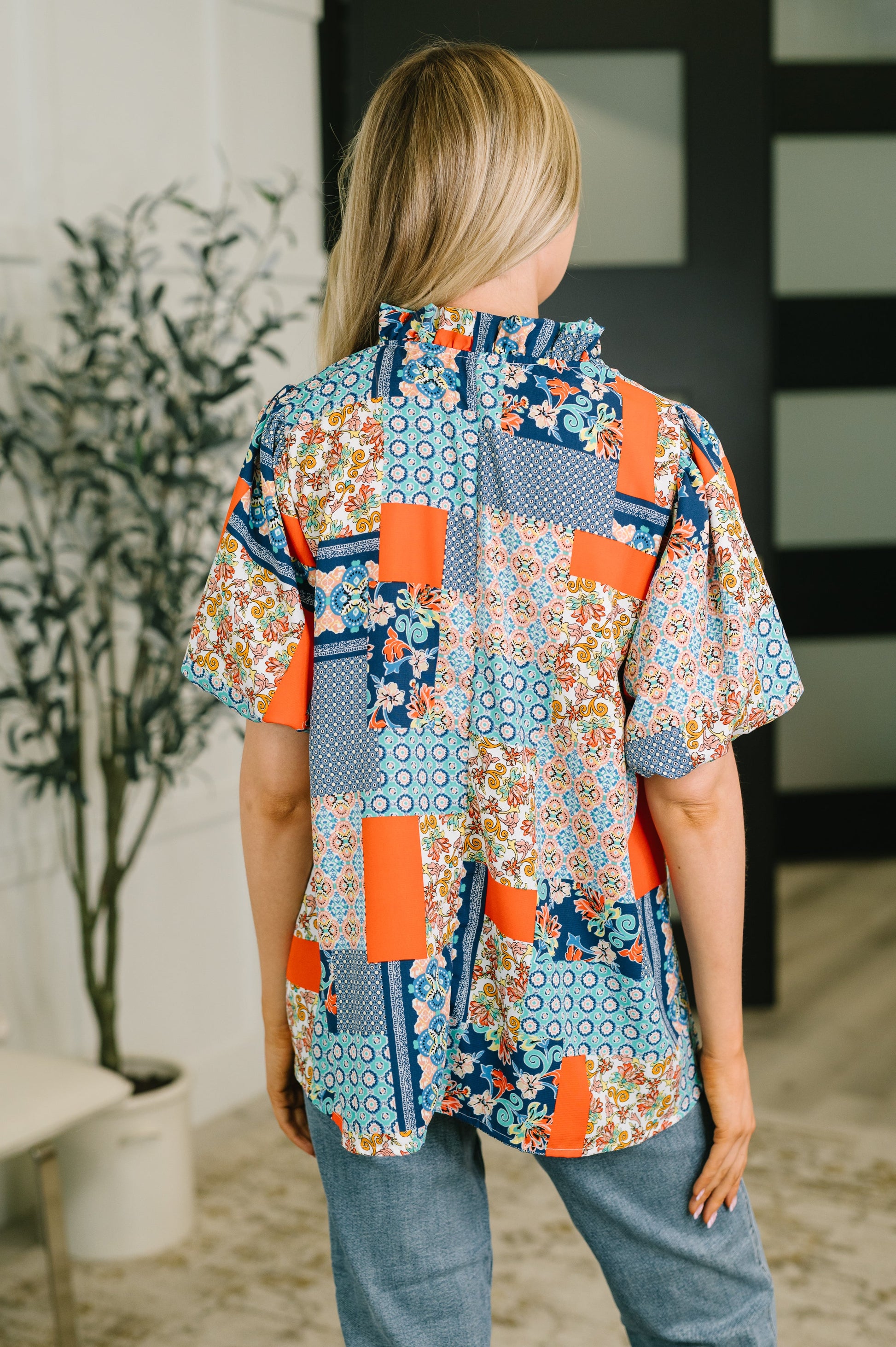 A woman viewed from behind wearing a short-sleeve patchwork button-up shirt with a colorful floral and geometric print in orange, blue, beige, and white, paired with blue jeans in an indoor setting.