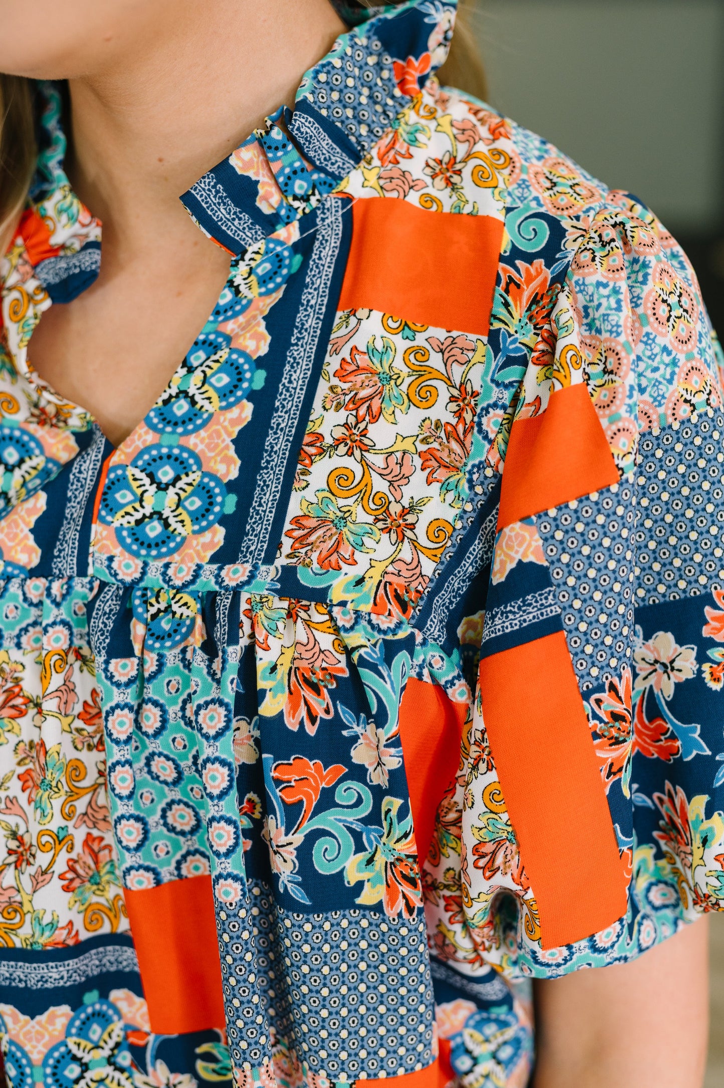 A close-up of a woman wearing a short-sleeve patchwork blouse with a mix of floral, paisley, and solid orange panels on a white background.