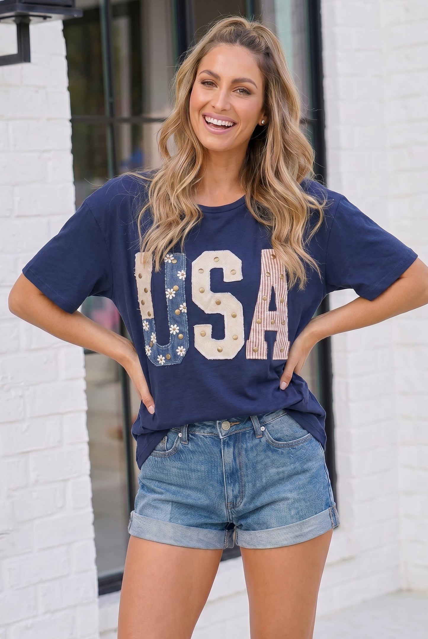 Woman wearing a navy blue 'USA' t-shirt and denim shorts standing against a white brick wall.