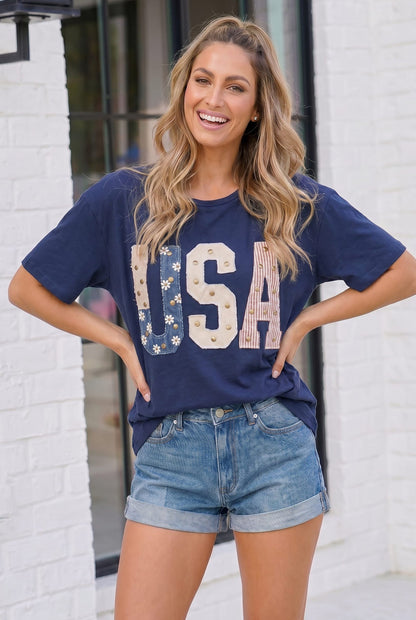 Woman wearing a navy blue 'USA' t-shirt and denim shorts standing against a white brick wall.