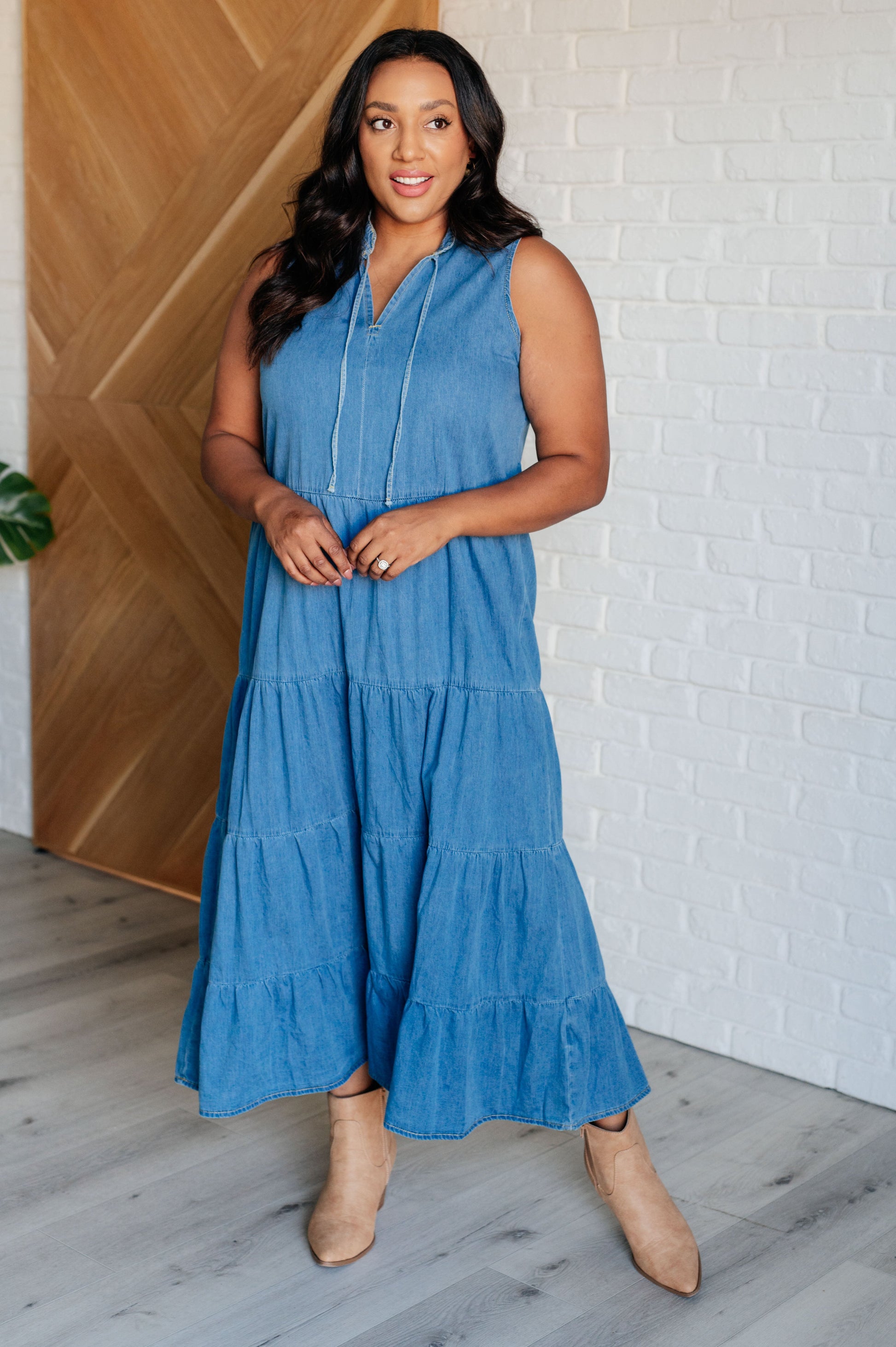 Woman wearing a blue sleeveless dress standing in a room with wooden flooring and a white brick wall.