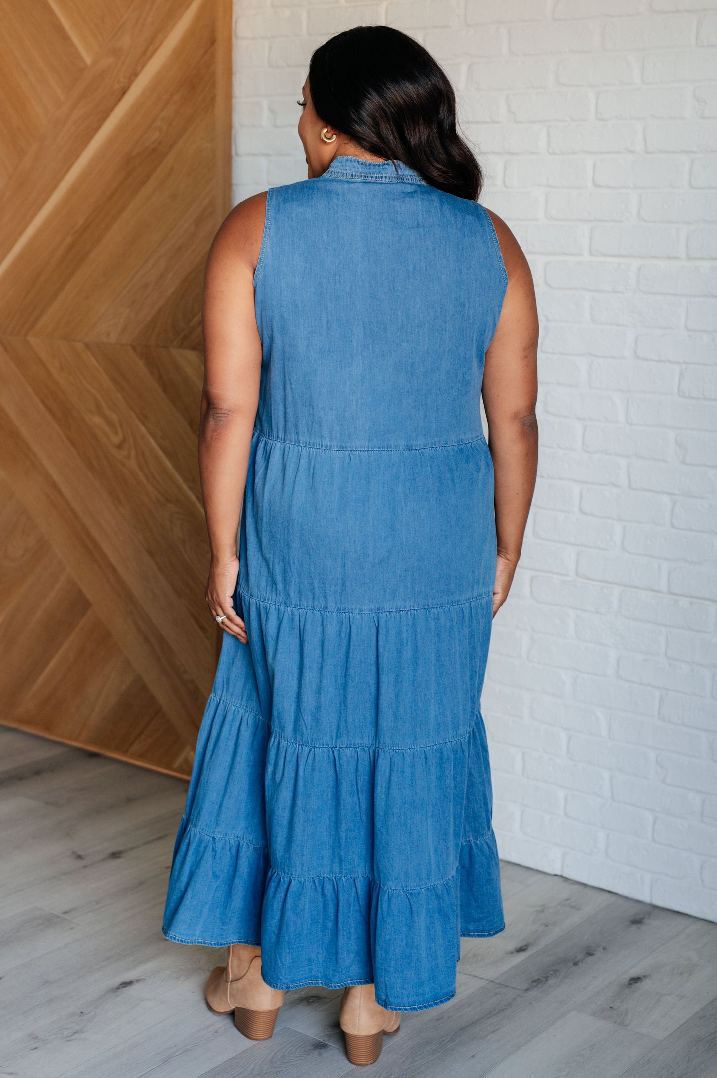 Woman wearing a blue dress standing against a wooden and white brick wall.