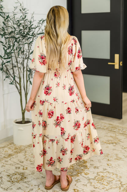 Woman wearing a floral dress standing in a room with a plant and door in the background.
