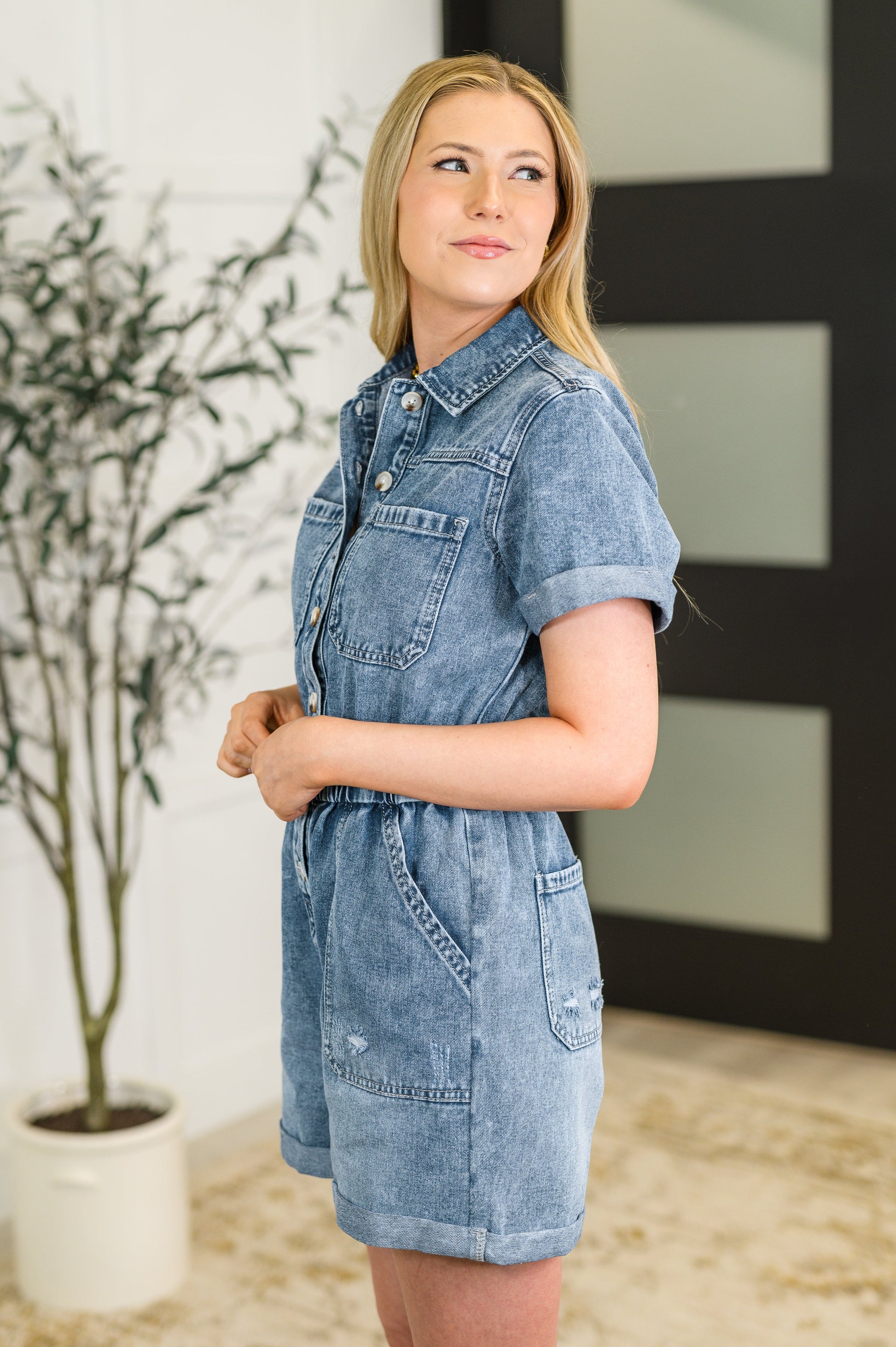 Woman wearing a denim dress standing indoors with a plant in the background