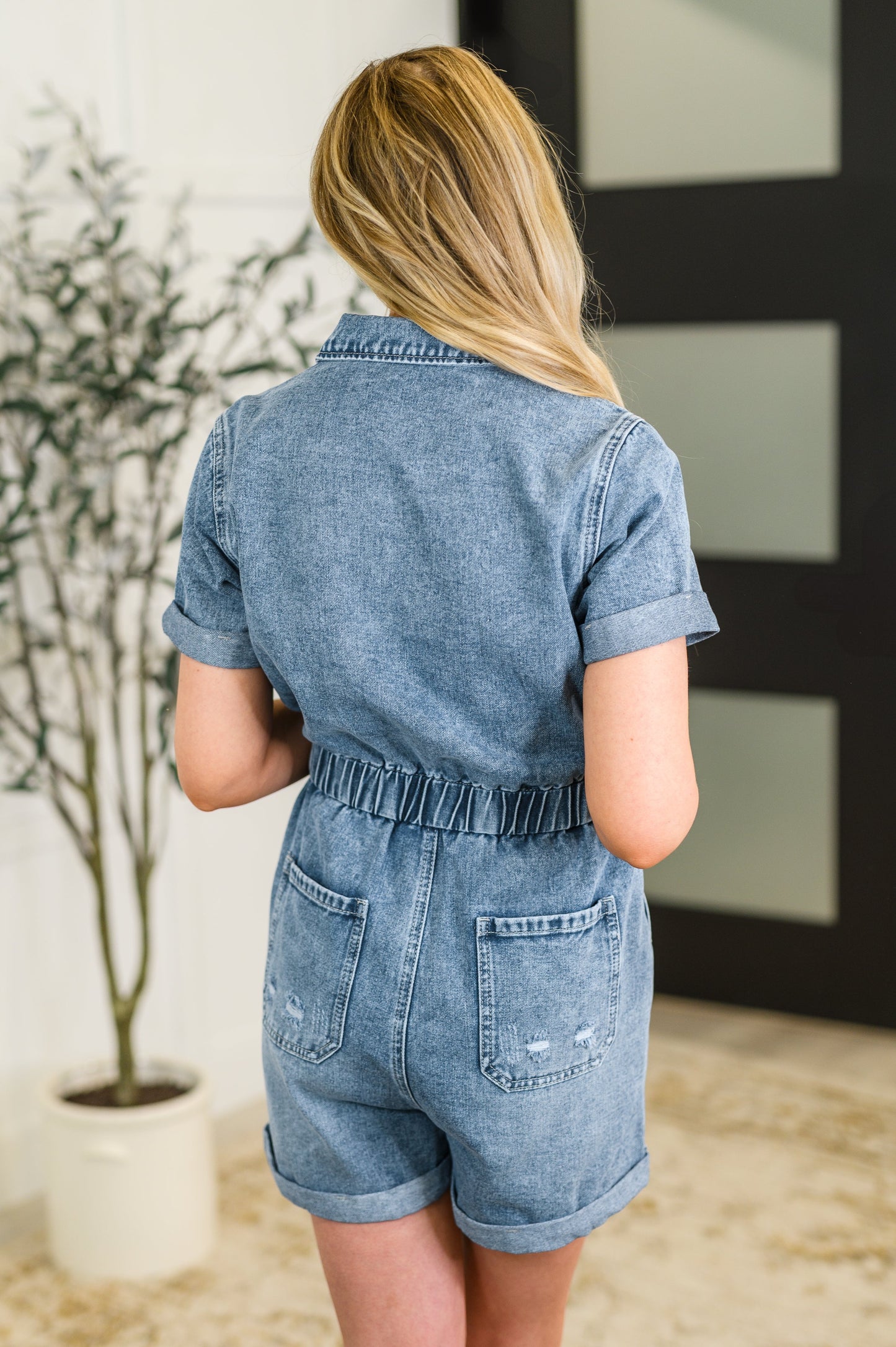 Person wearing a denim romper standing in a room with a plant and framed picture.