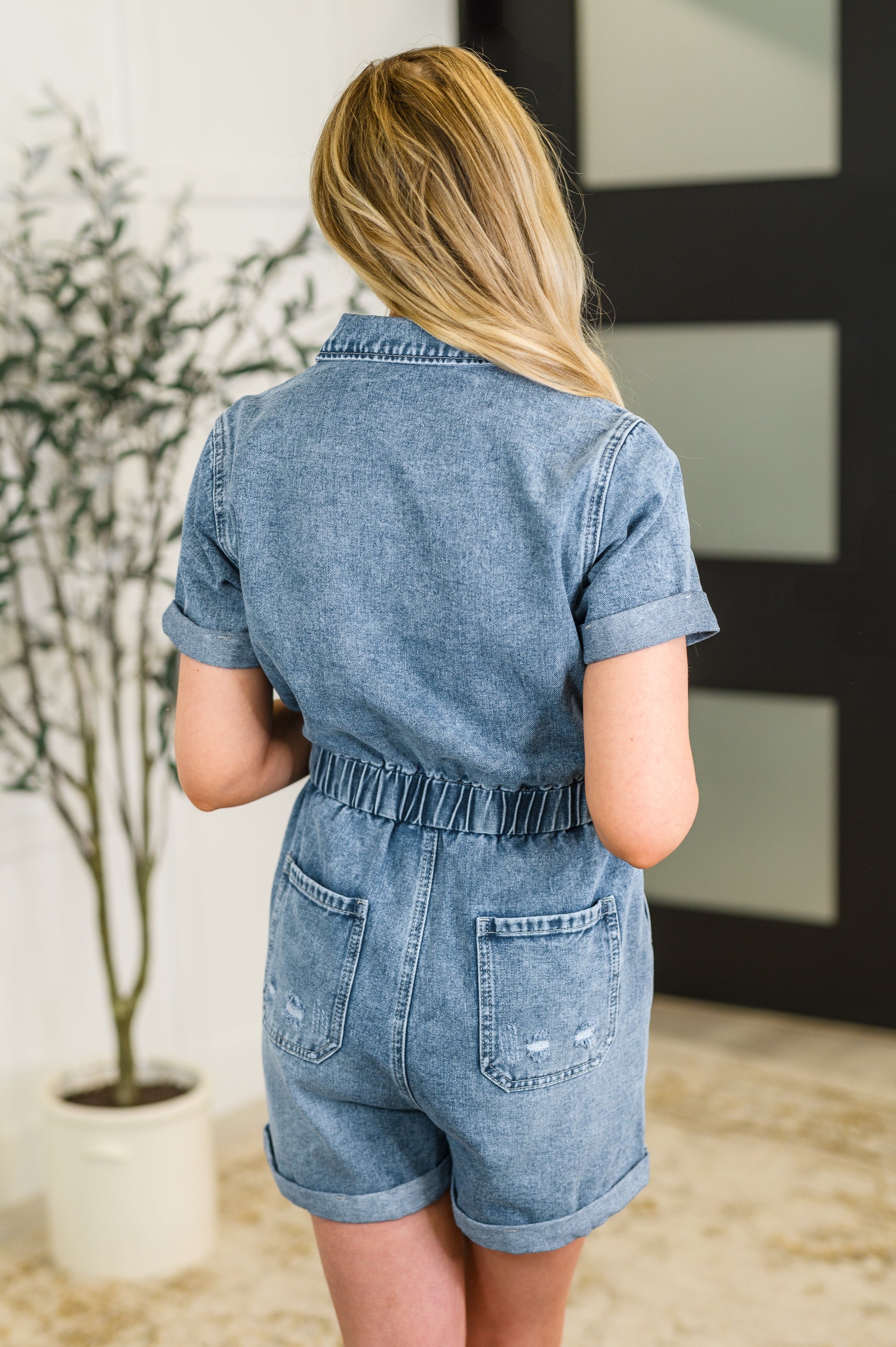 Person wearing a denim romper standing in a room with a plant and framed picture.
